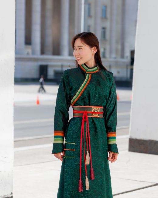 Asian woman in traditional green robe standing outdoors in urban setting with architectural background.
