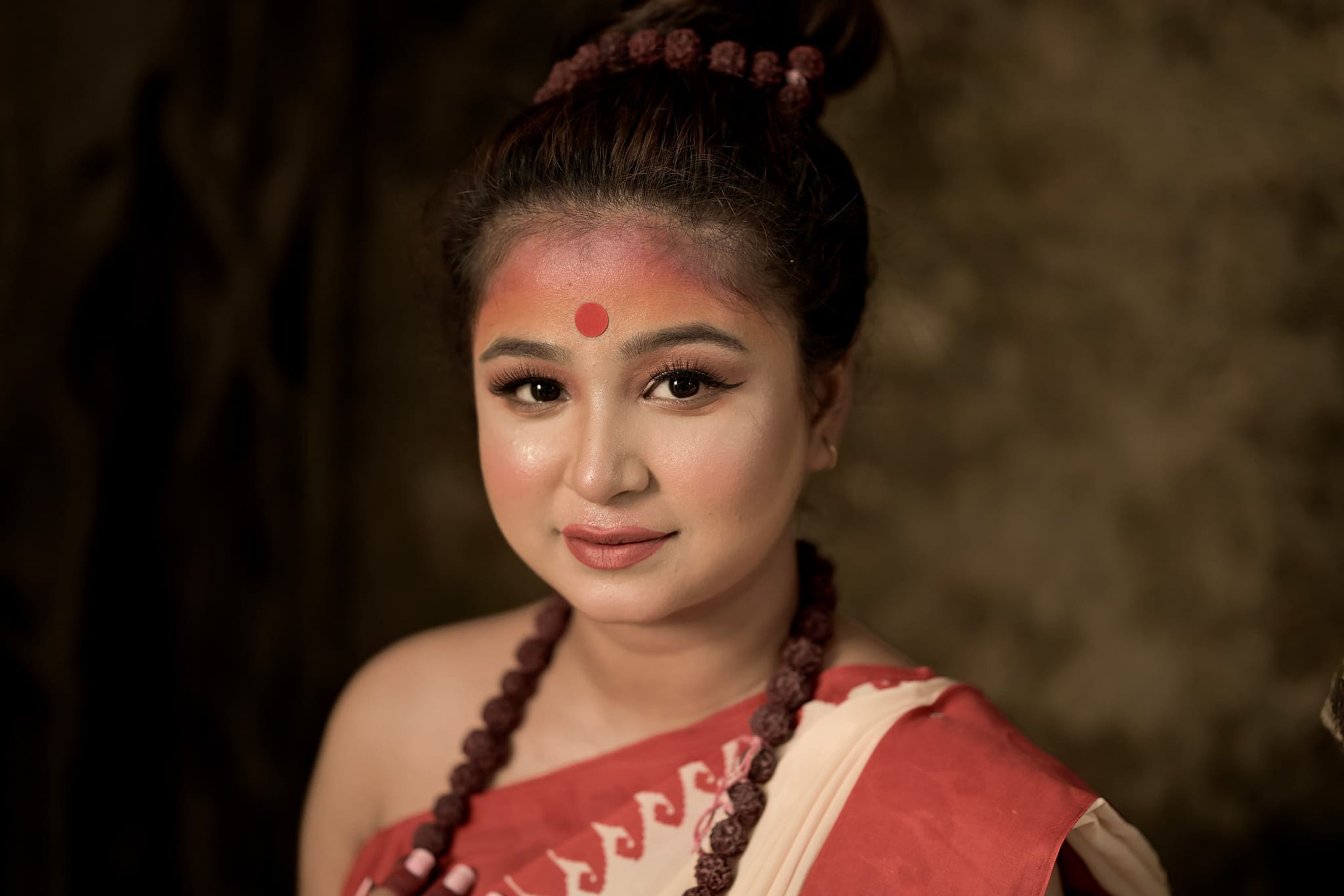 Close-up portrait of an adult woman with traditional red bindi and beads, wearing a red and white saree.