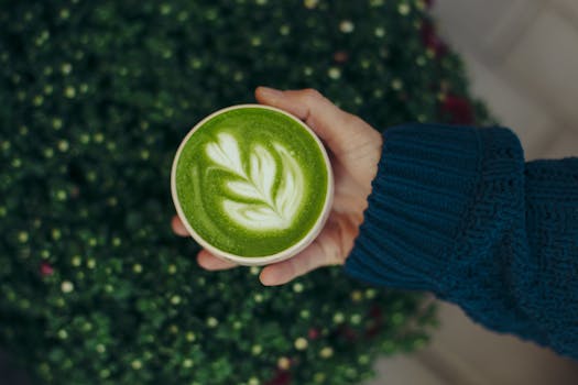 A close-up of a matcha latte with latte art, held by a hand in a cozy knitted sweater, against a blurred green background.