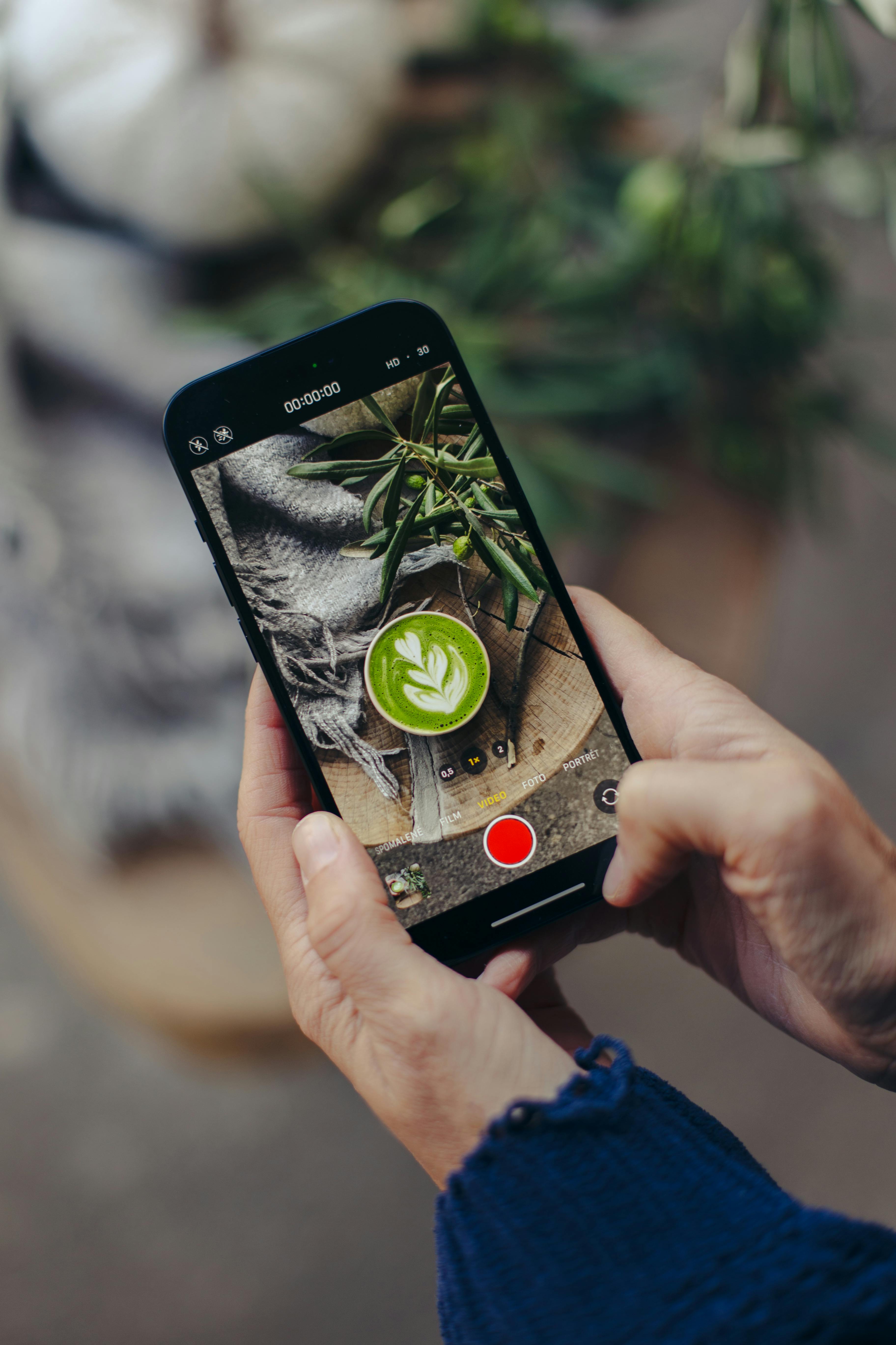 Hands using smartphone to photograph a matcha latte with autumn leaves in Sabinov, Slovakia.