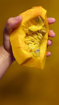 A close-up of a hand holding a fresh pumpkin slice with seeds on a yellow background.