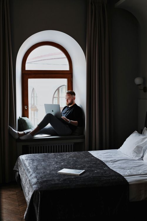 Free A man sits comfortably on a window ledge using a laptop in a cozy hotel room. Stock Photo
