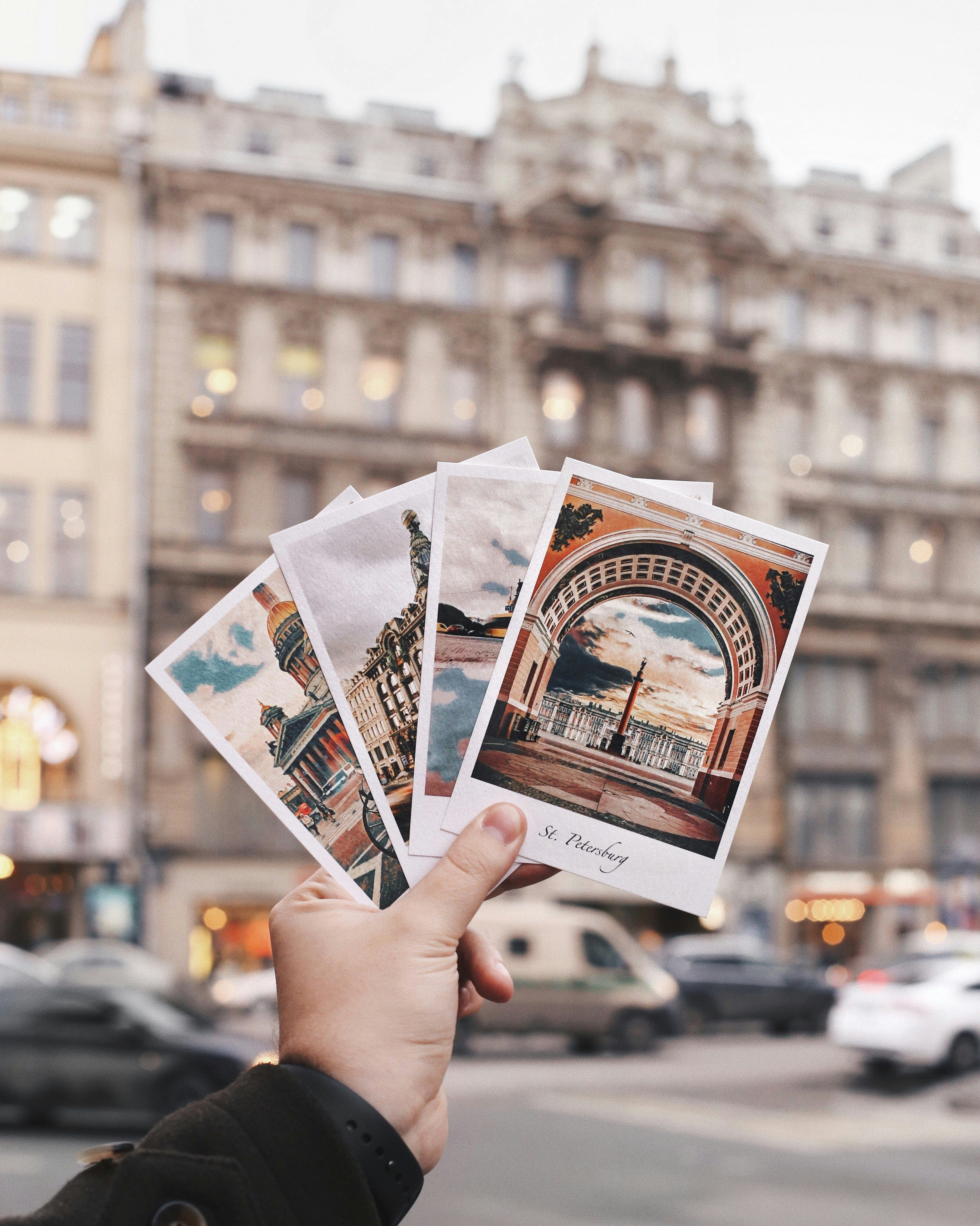 A hand holds postcards in front of a historic building in Saint Petersburg, Russia.