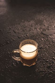 一个盛着泡沫咖啡的透明玻璃杯，放在潮湿阴暗的桌面上，营造出一种忧郁而诱人的氛围