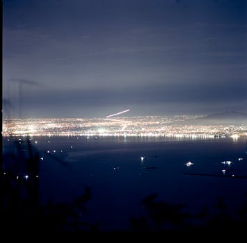 A breathtaking aerial night view of a coastal city illuminated with vibrant lights under a clear sky.
