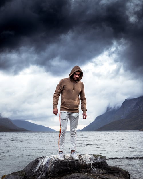 Kostenlos Mann, Der Braunes Sweatshirt Und Weiße Jogginghose Trägt, Die Auf Felsen Nahe Körper Des Wassers Stehen Stock-Foto