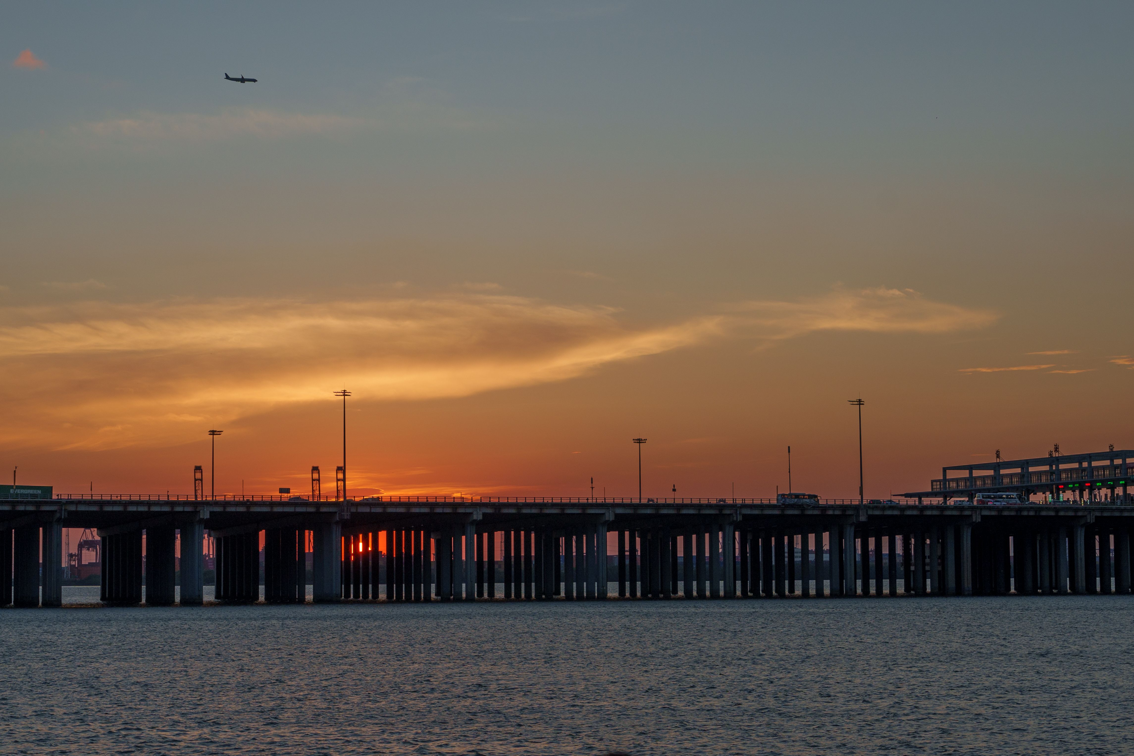 An airplane flying over a bridge silhouetted against a vibrant sunset sky.