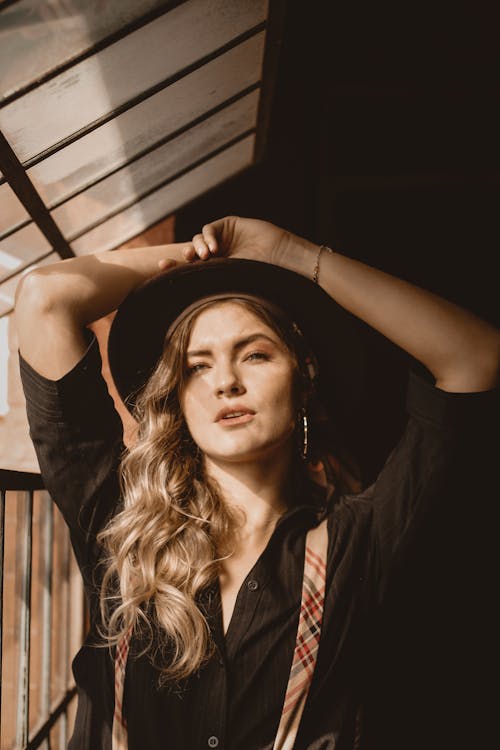 Photo of Woman in Black Top and Hat Posing by Window with Her Hands Over Her Head