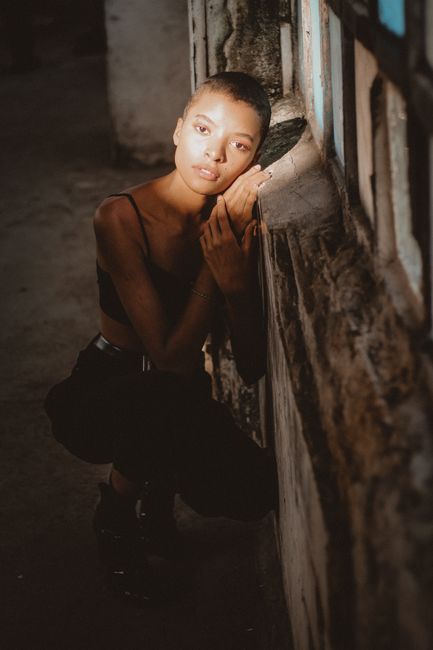 Model posing in casual attire by a textured wall in soft lighting.