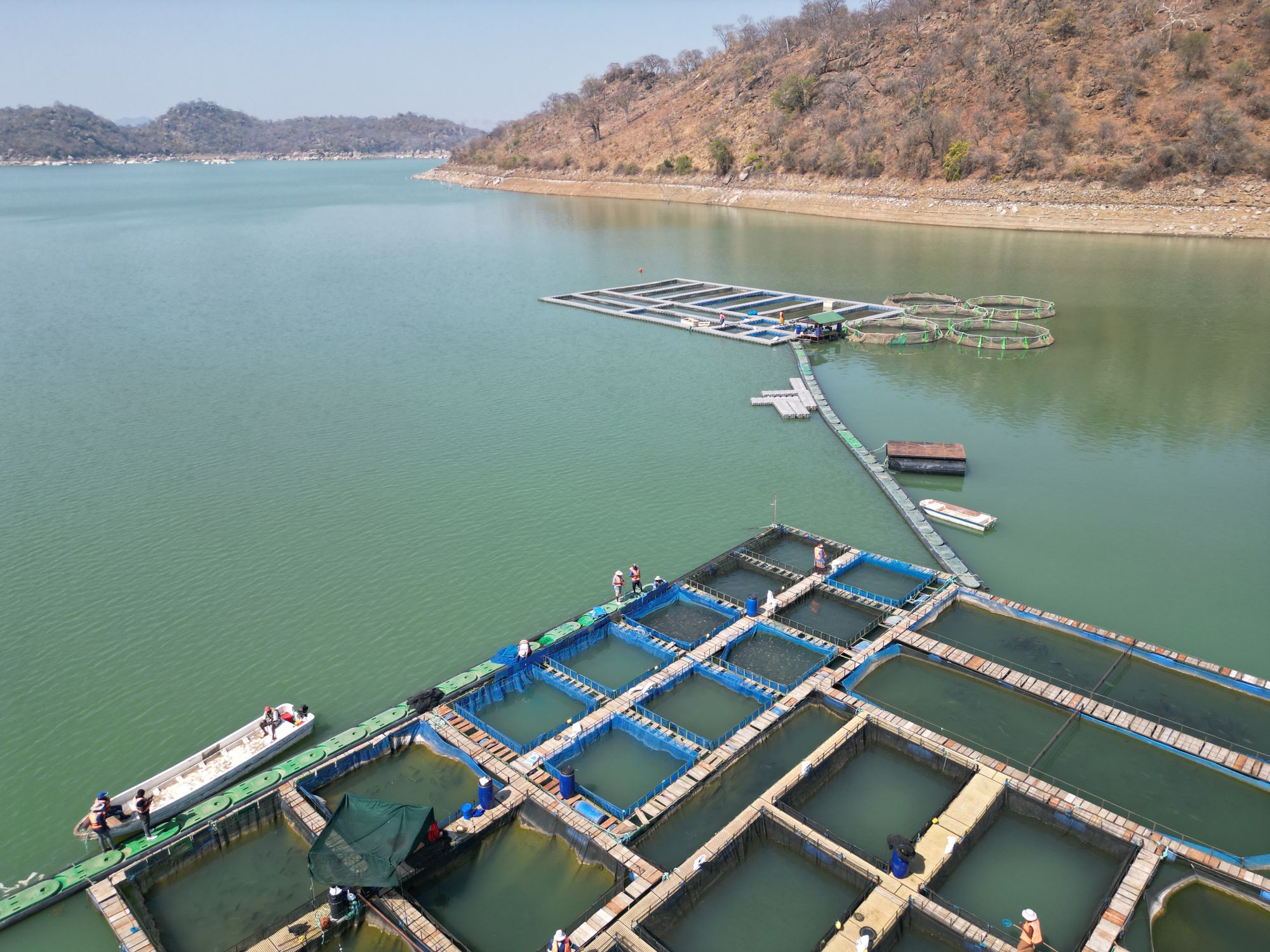 Aerial view of a traditional fish farm on a serene lake with clear skies and surrounding hills.