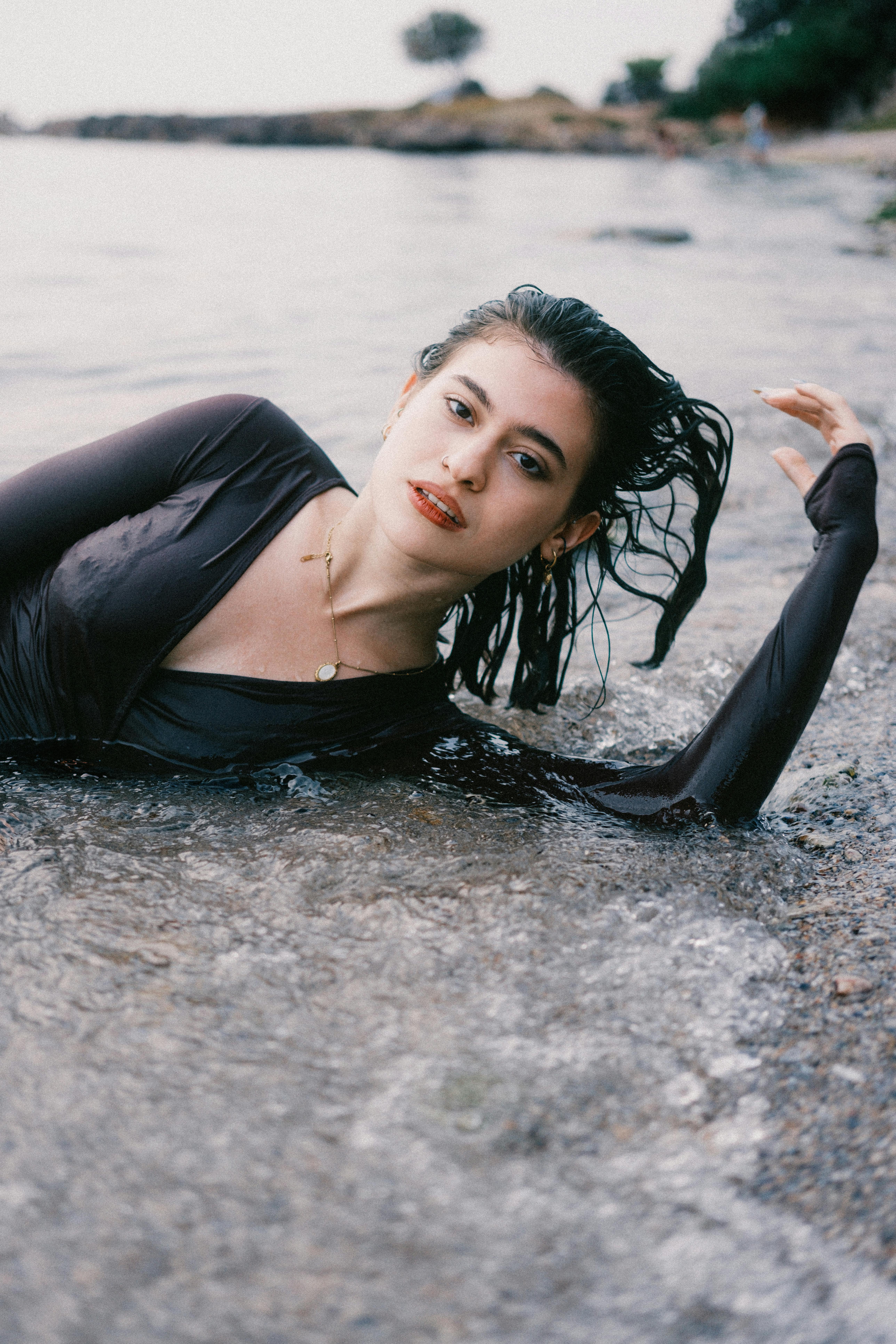 A woman in black dress lies elegantly on water's edge, natural setting.