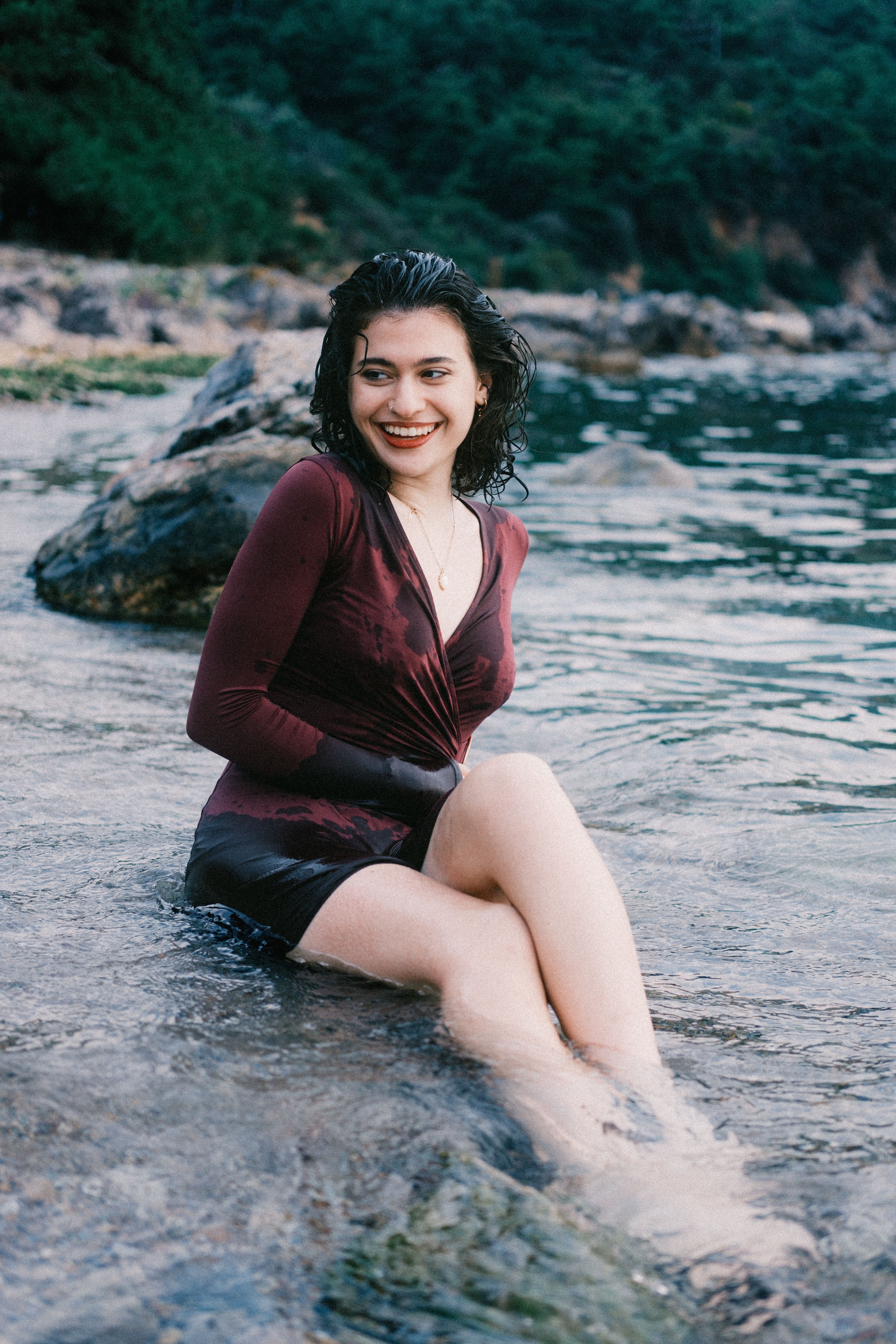 Woman sitting in water at the edge of a tranquil rocky shore, smiling.