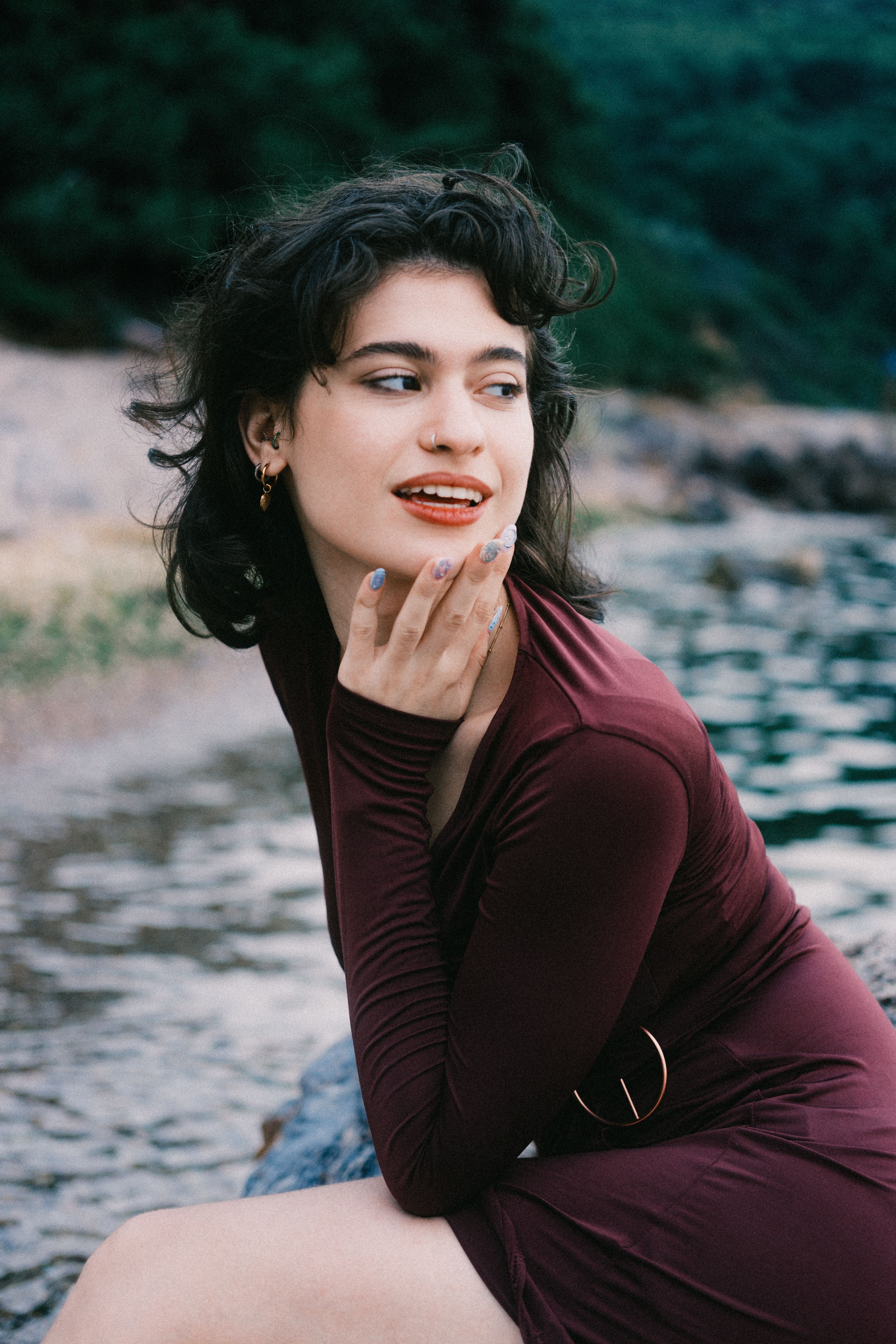 Stylish woman in maroon dress enjoying a moment by a peaceful lake. Captivating smile and relaxed pose.