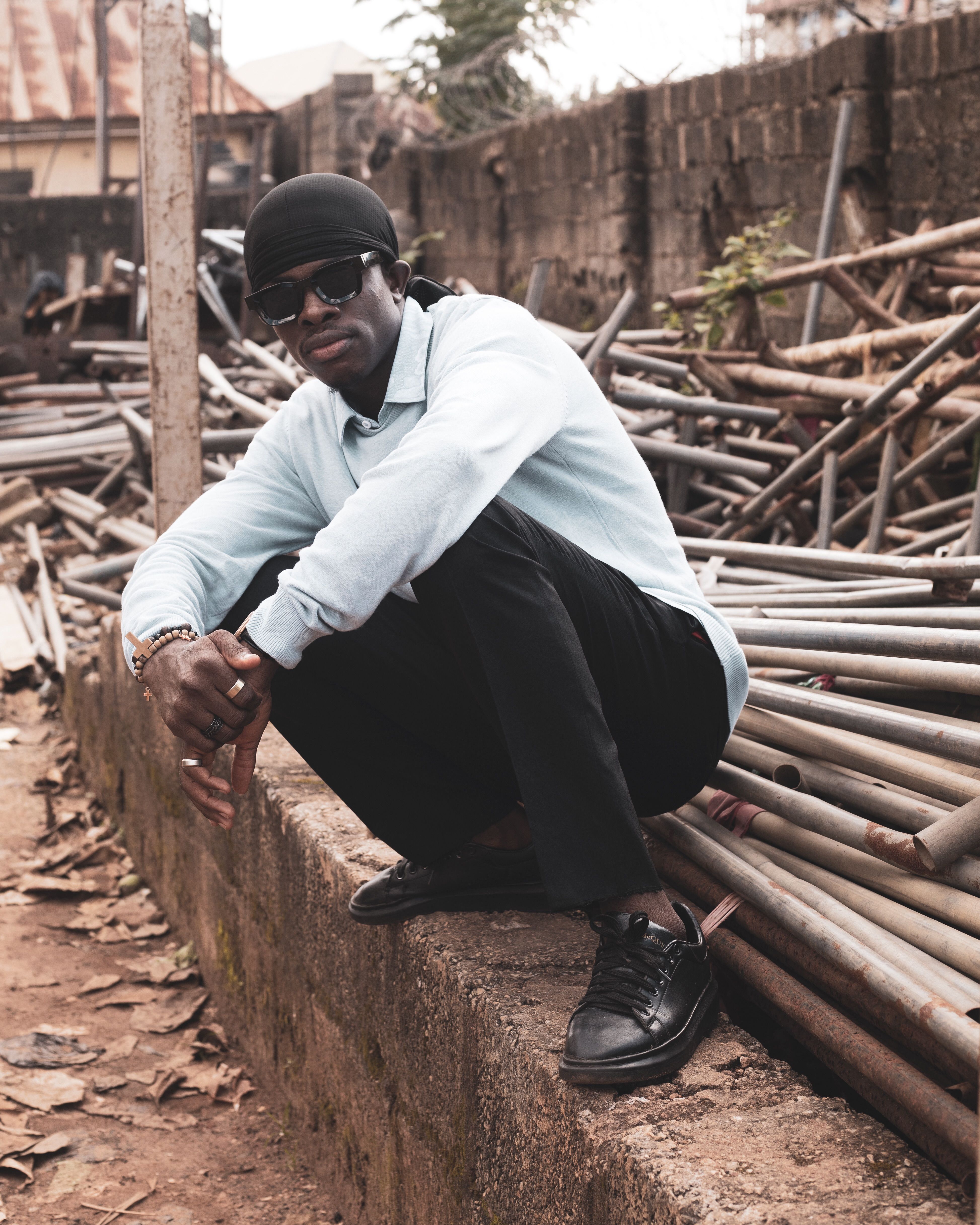 Man in casual attire squatting beside metal pipes in an outdoor industrial setting.