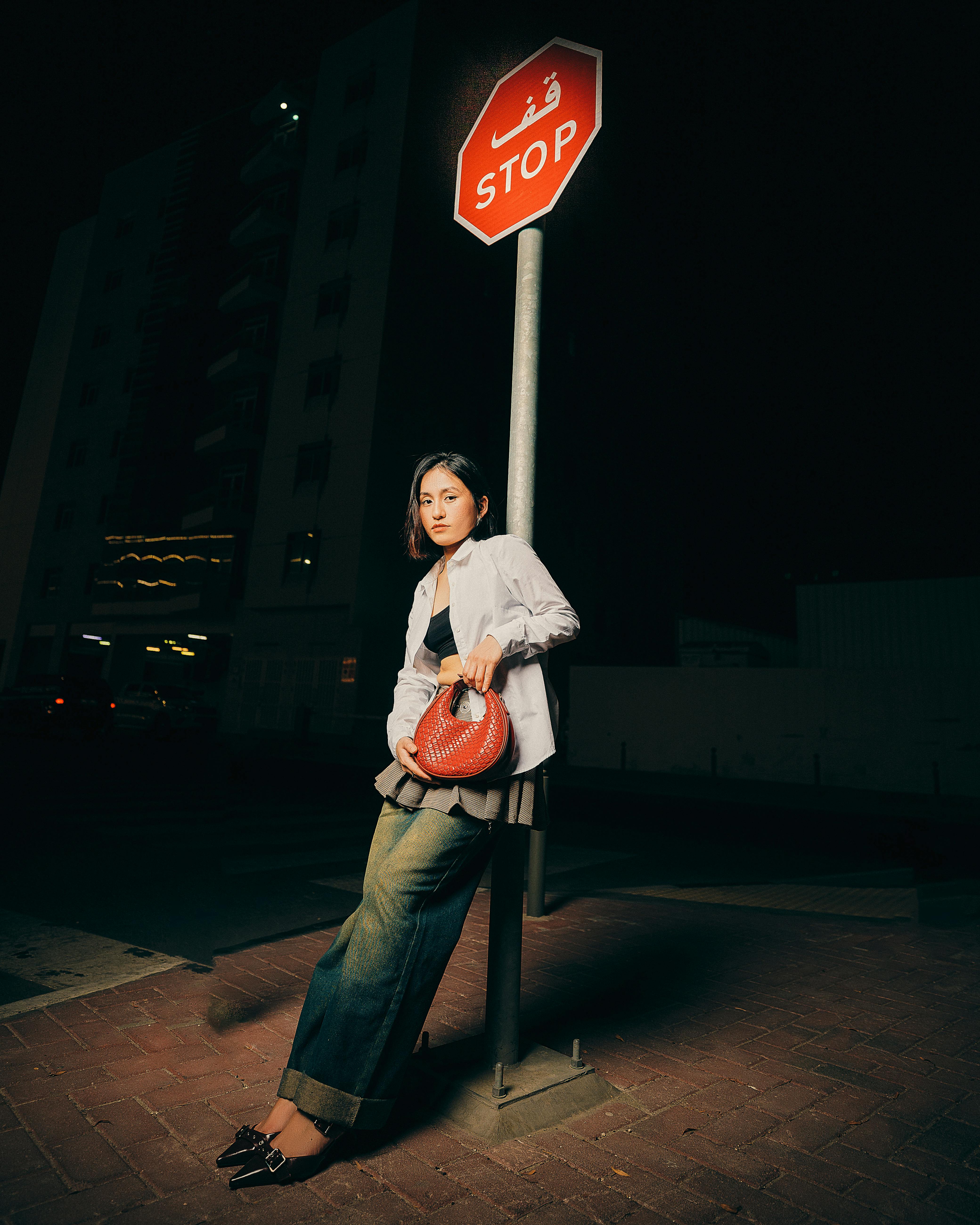 Stylish woman holding a red bag by a stop sign in an urban setting at night.