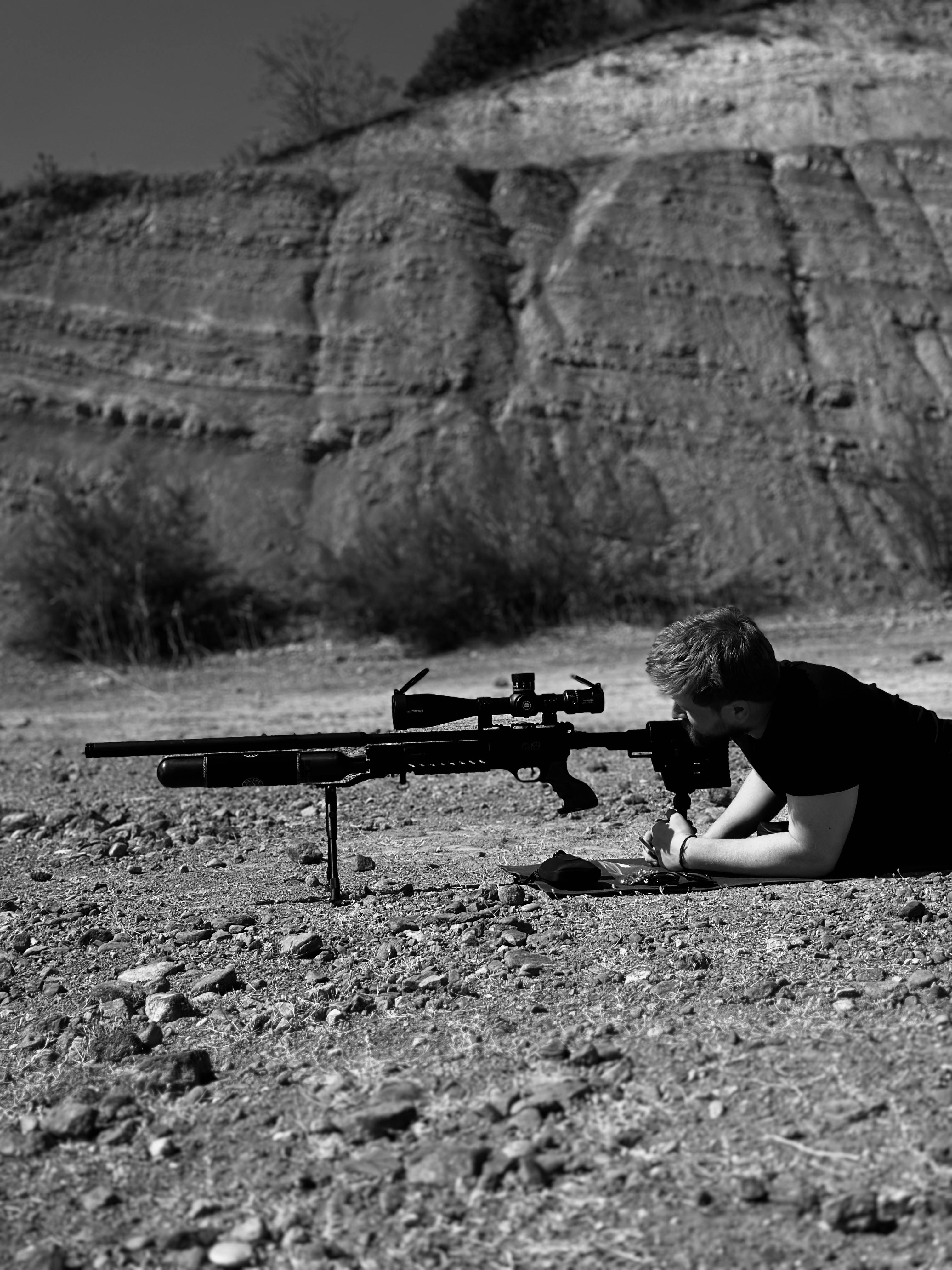 Person lying prone with rifle in rocky desert setting. Black and white photo.