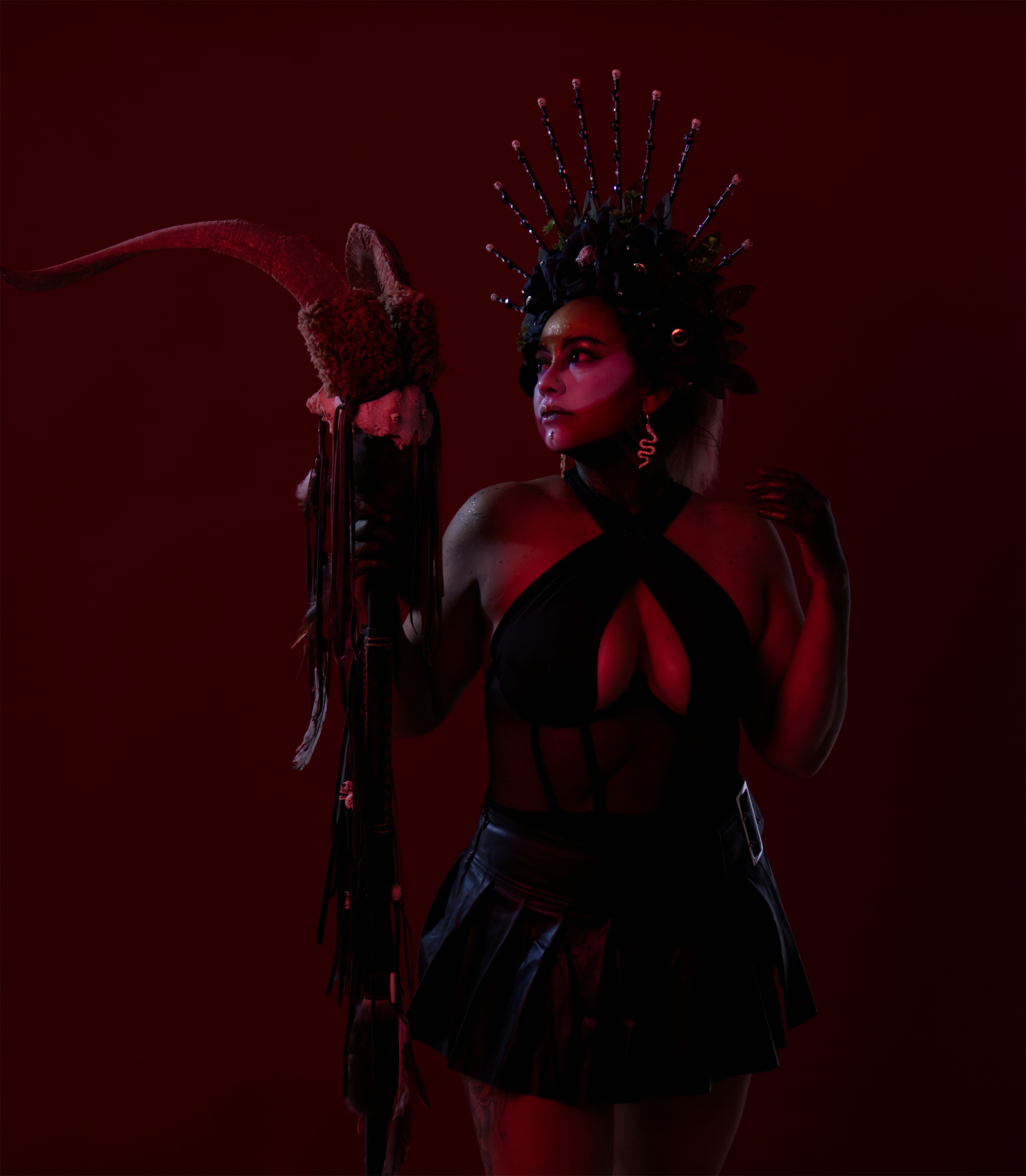 A woman in dark, dramatic attire and a striking headdress holds a mystical staff against a red backdrop.