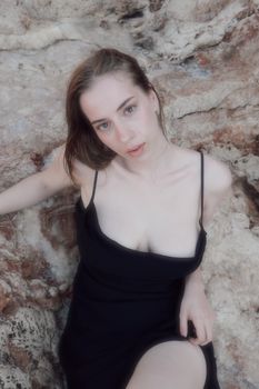 Fashionable woman in black dress sitting on rocky terrain, serene expression.