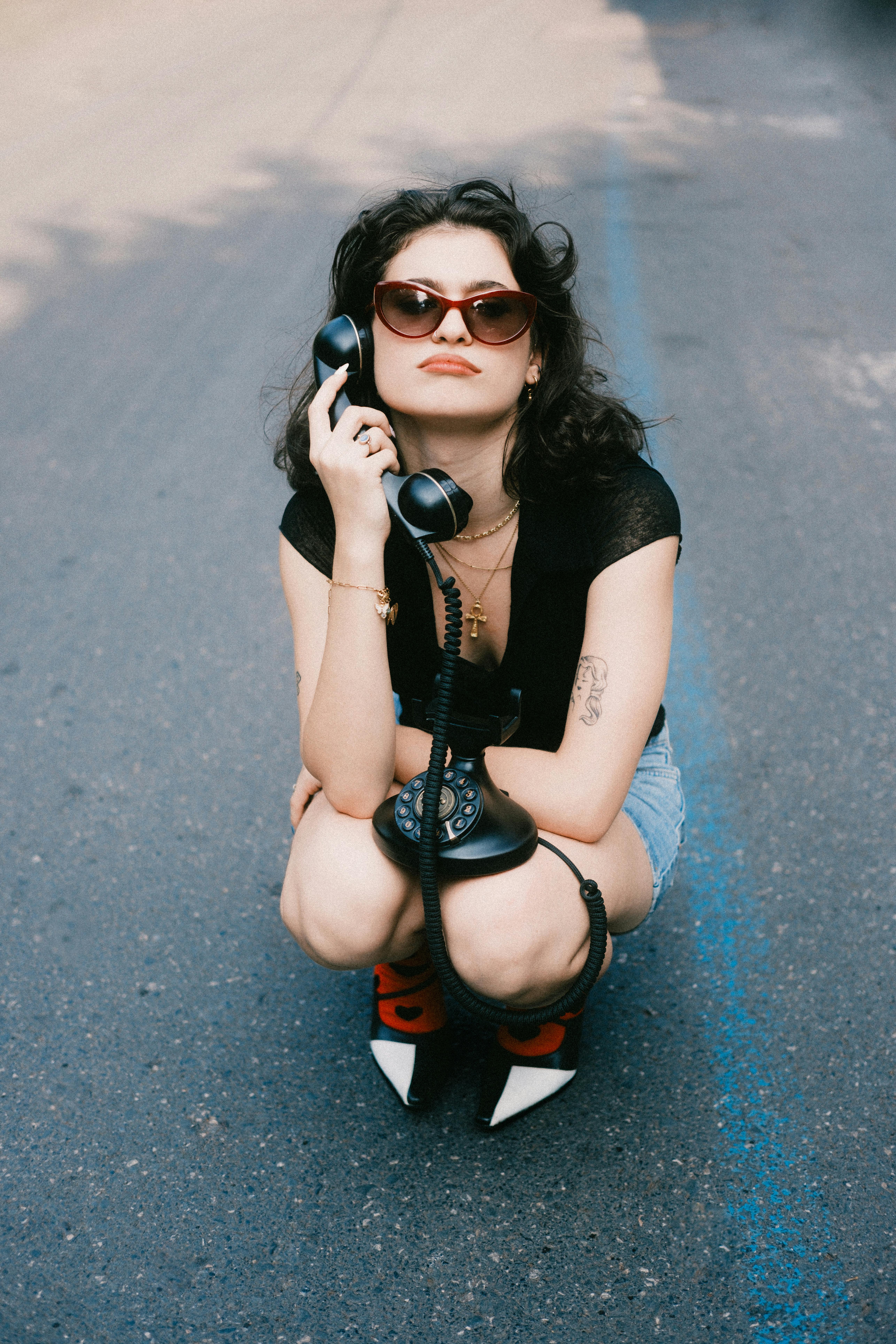 Stylish woman squatting on the street with a retro telephone and sunglasses.