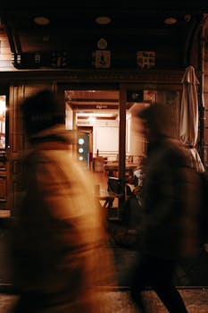 Motion blur of people walking past a warmly lit cafe at night, creating a dynamic urban scene.