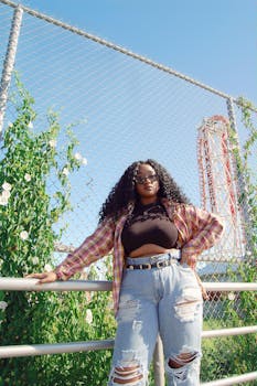 Fashionable woman with curly hair and sunglasses posing stylishly outdoors by a fence.
