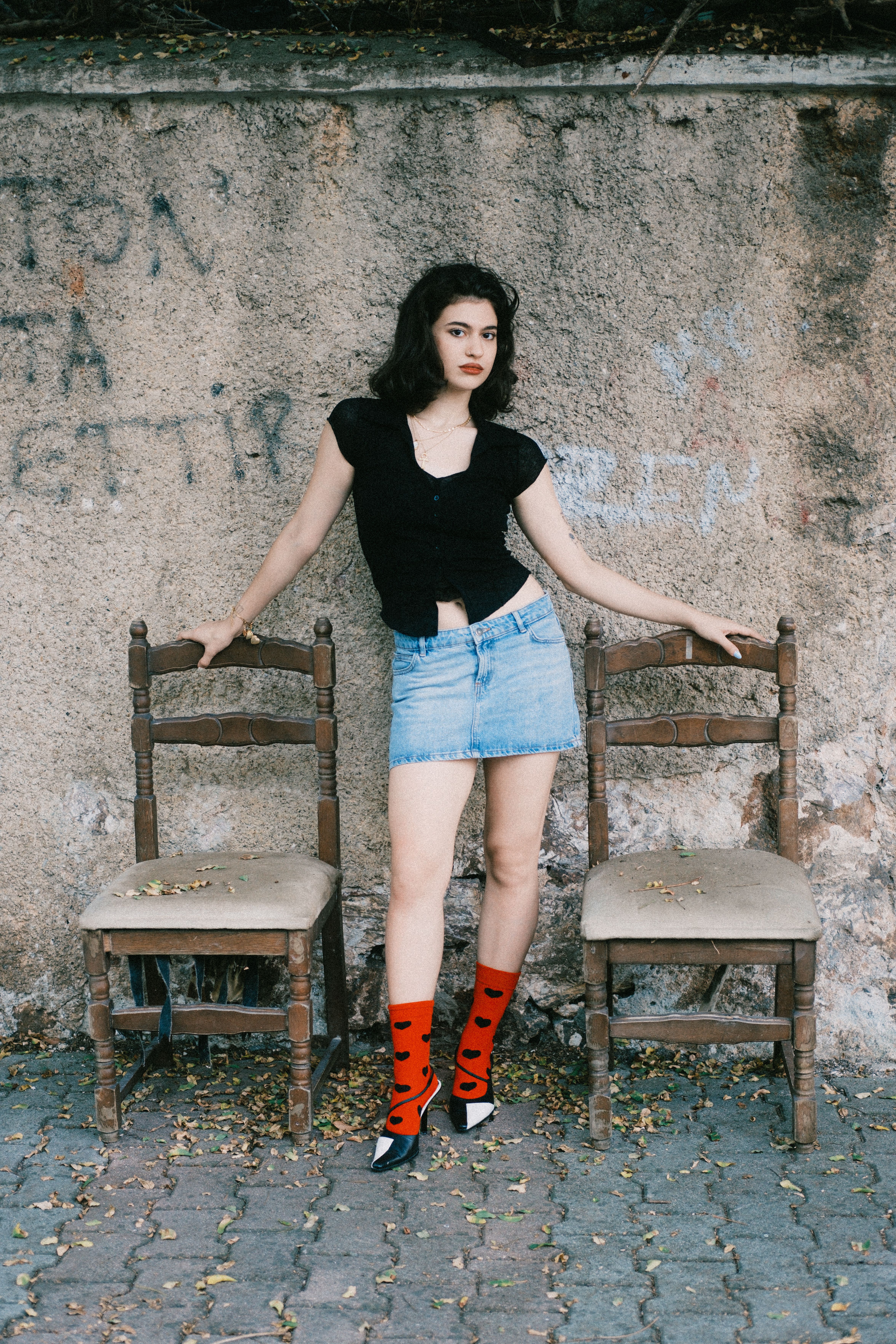 Fashionable young woman posing outdoors in a chic outfit with vintage chairs.