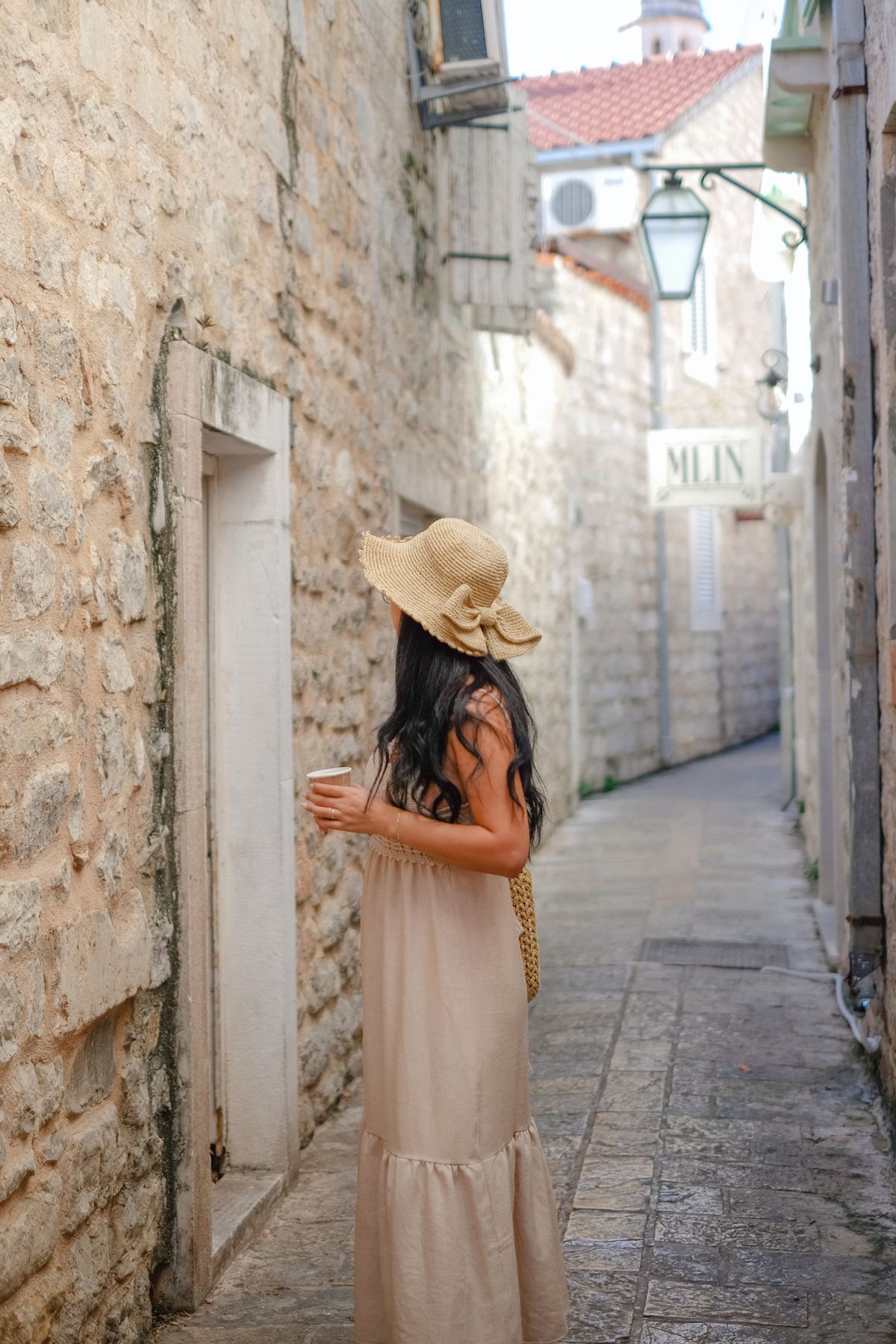 A woman exploring a scenic narrow European street — the raffia bag and linen ease of a European summer