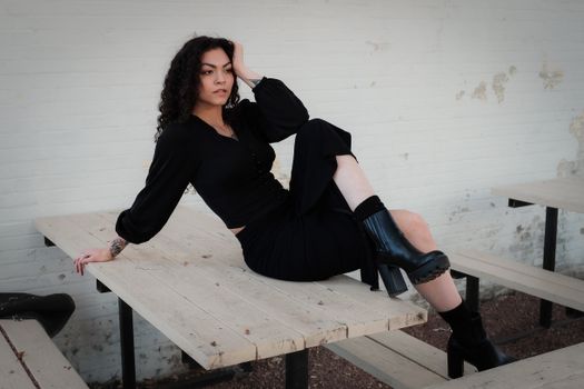 Fashionable woman dressed in black sitting confidently on an outdoor bench against a light wall.