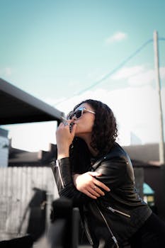 Fashionable woman wearing sunglasses in a leather jacket posing confidently against a city backdrop.