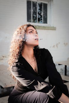 A contemplative woman smoking outside, highlighted by natural light against a rustic backdrop.