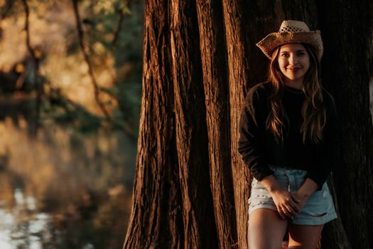 Portrait of a woman in a straw hat leaning against a tree by the water.