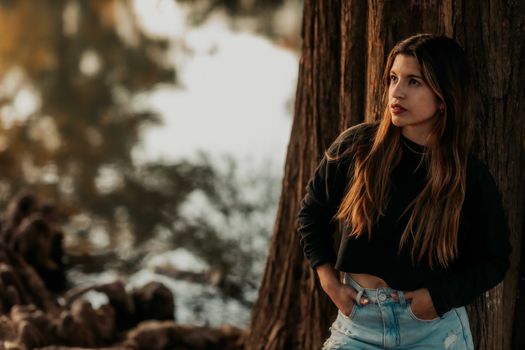 A young woman in a black top and jeans leans against a tree by a calm lake.