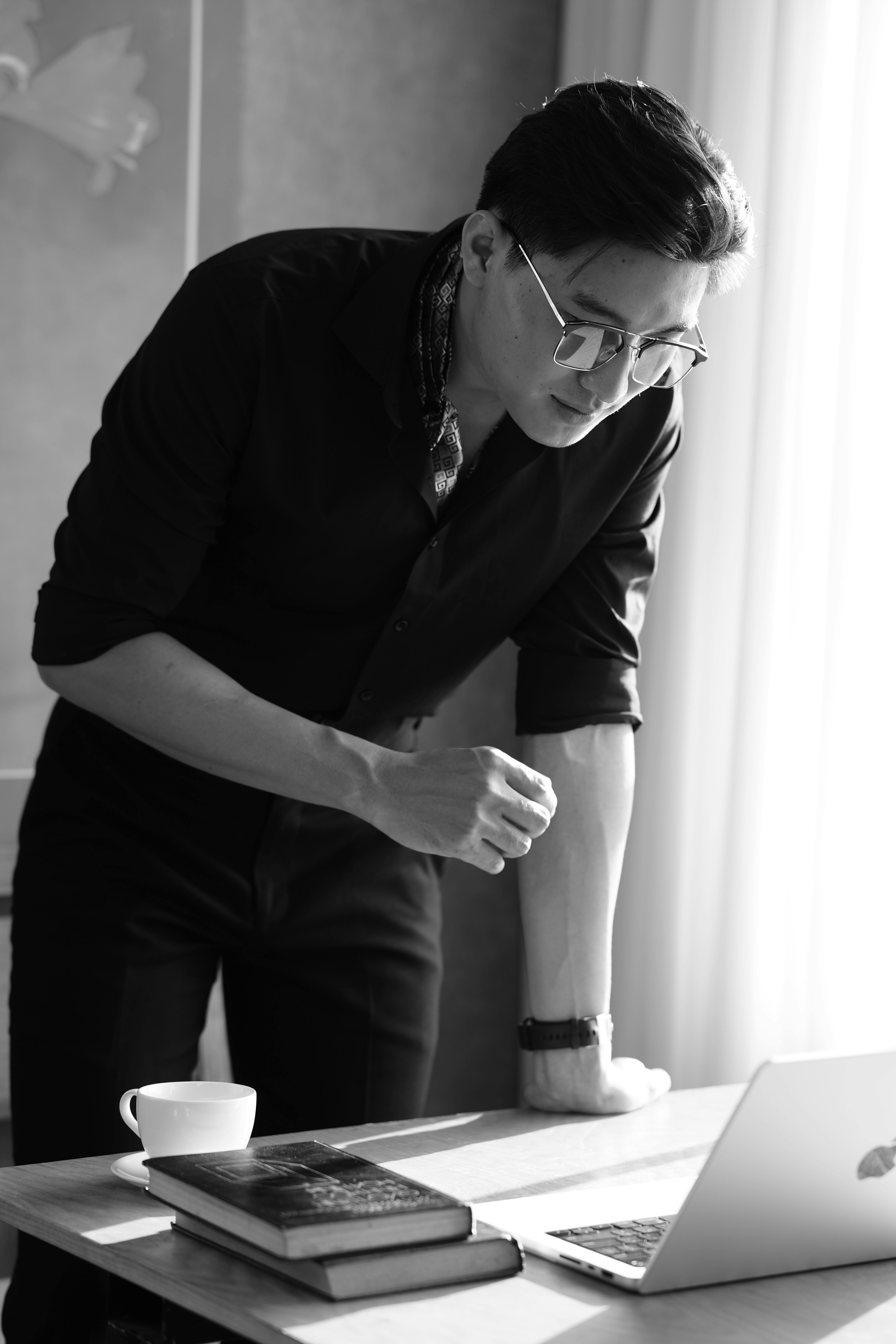 Focused businessman leaning over a desk with a laptop, books, and cup in a modern office space.