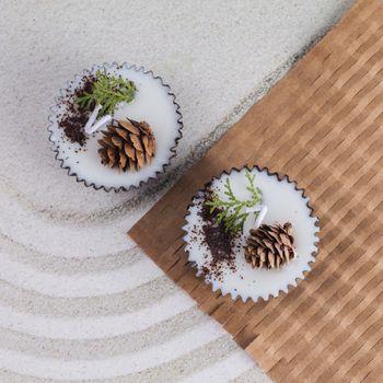 Top view of decorative candles with pinecones and greenery set on sand and paper, emphasizing natural aesthetics.