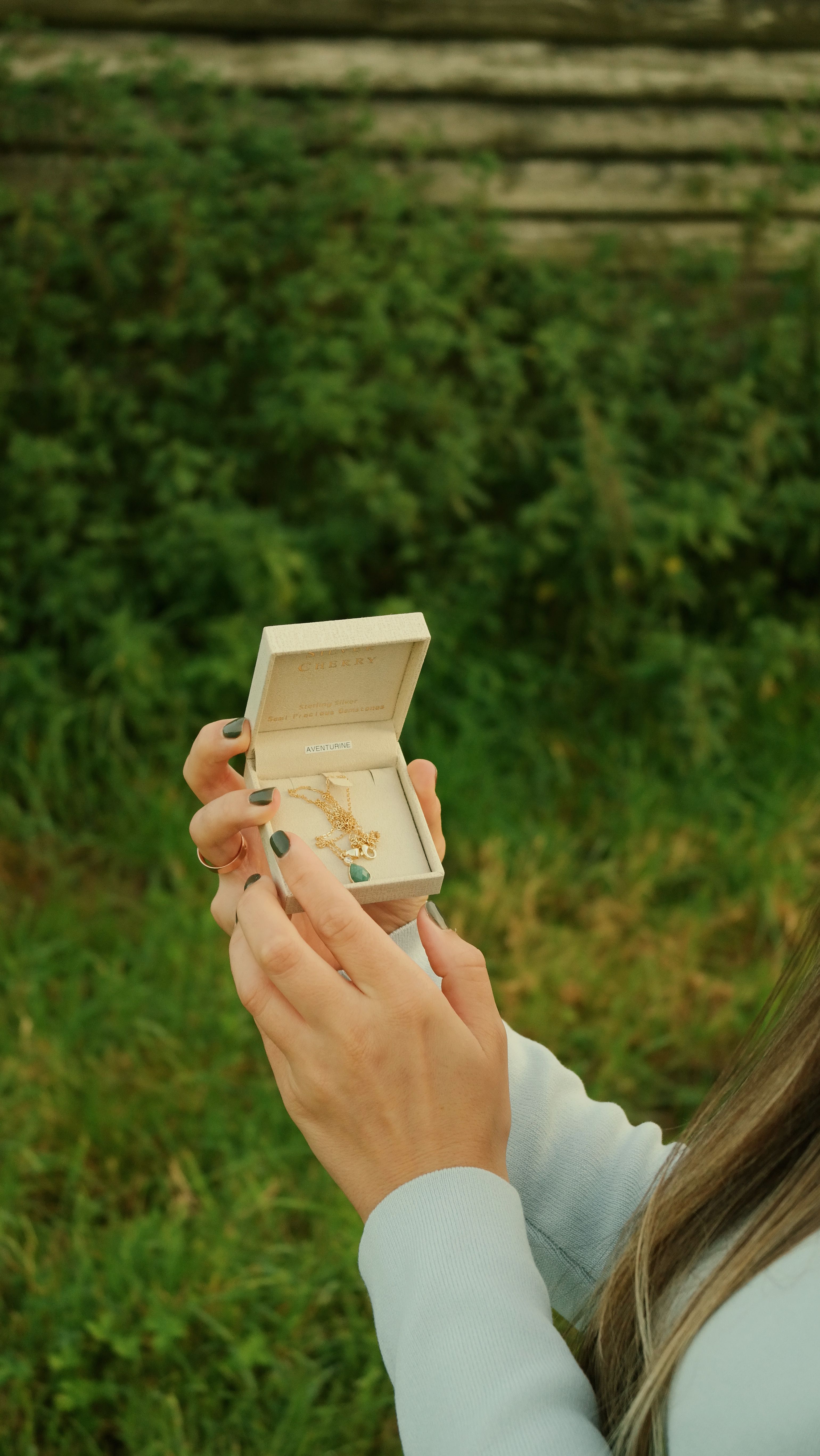 A woman's hands open a jewelry box displaying a necklace against a natural background.