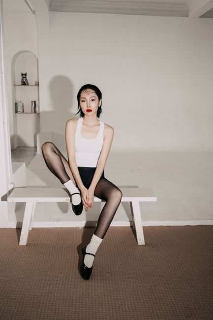 Artistic portrait of a woman in monochrome attire seated indoors in a minimalist style.