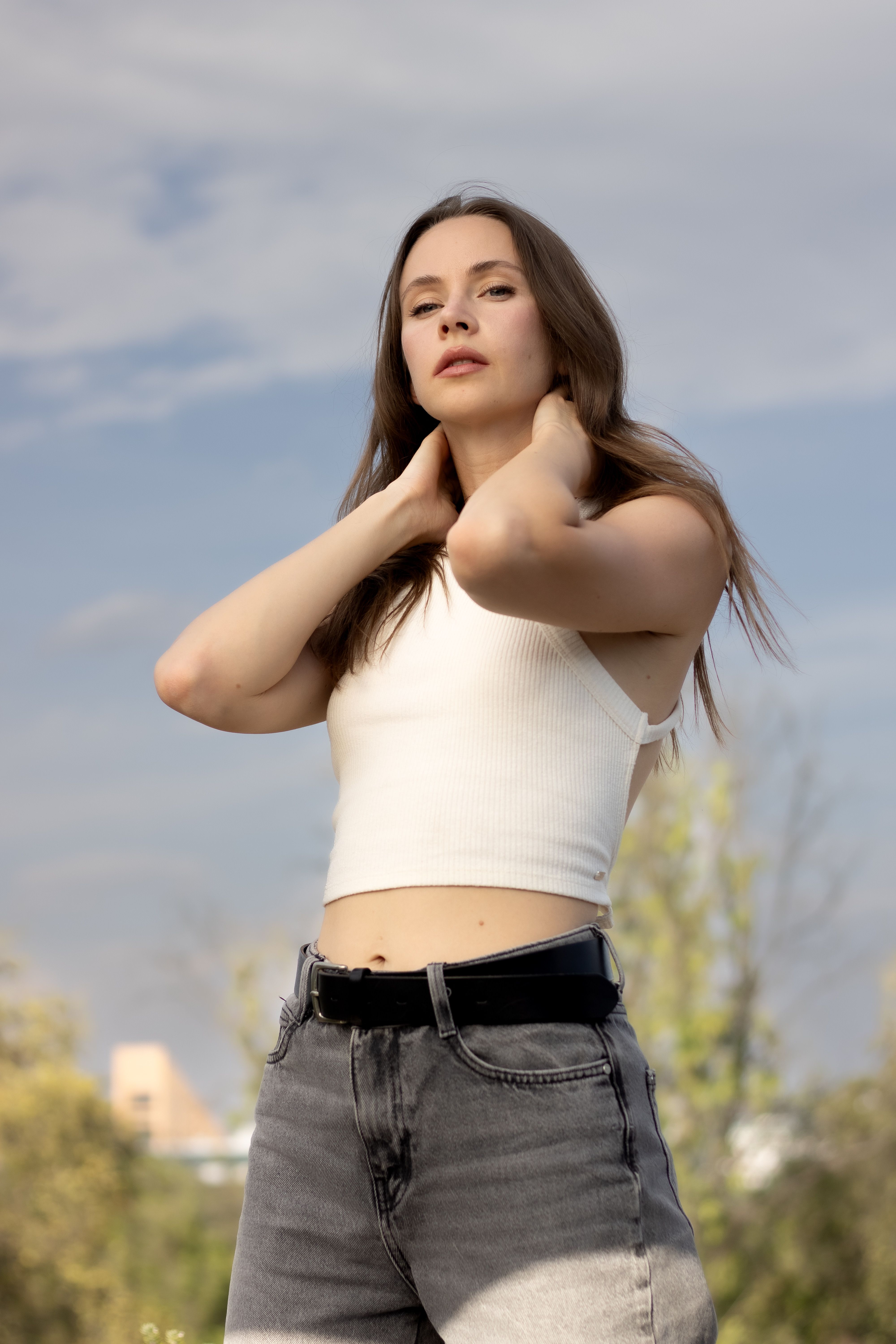 Young woman in white top posing outdoors with sky backdrop.