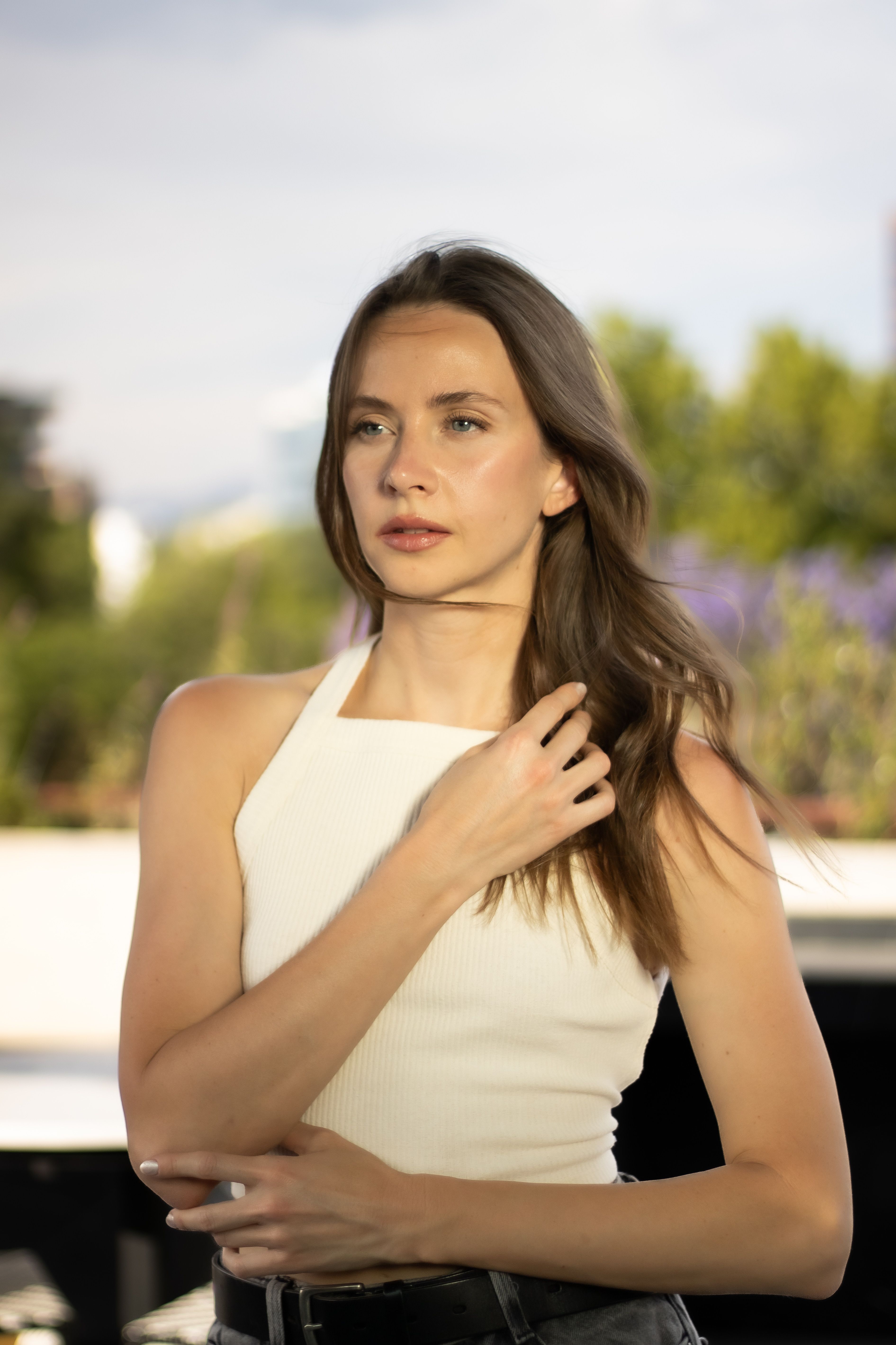 A young woman in a white top poses naturally outdoors against a blurred background.