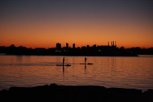 Free Two paddleboarders silhouetted against a vibrant sunset over a calm lake with city skyline. Stock Photo