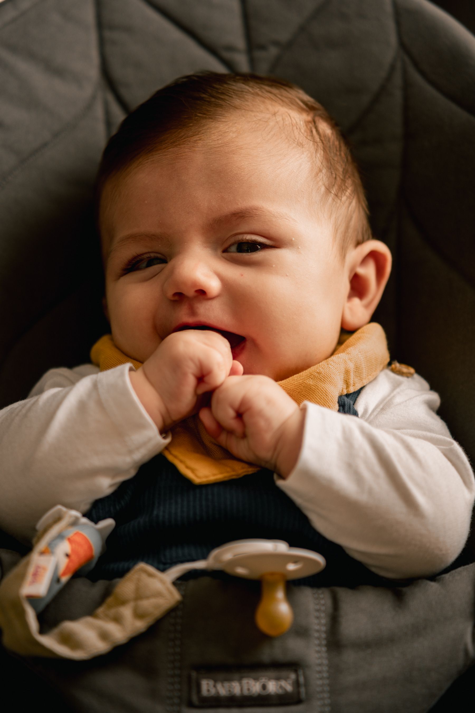 Charming portrait of a baby sitting in a car seat, chewing hands and looking curious.