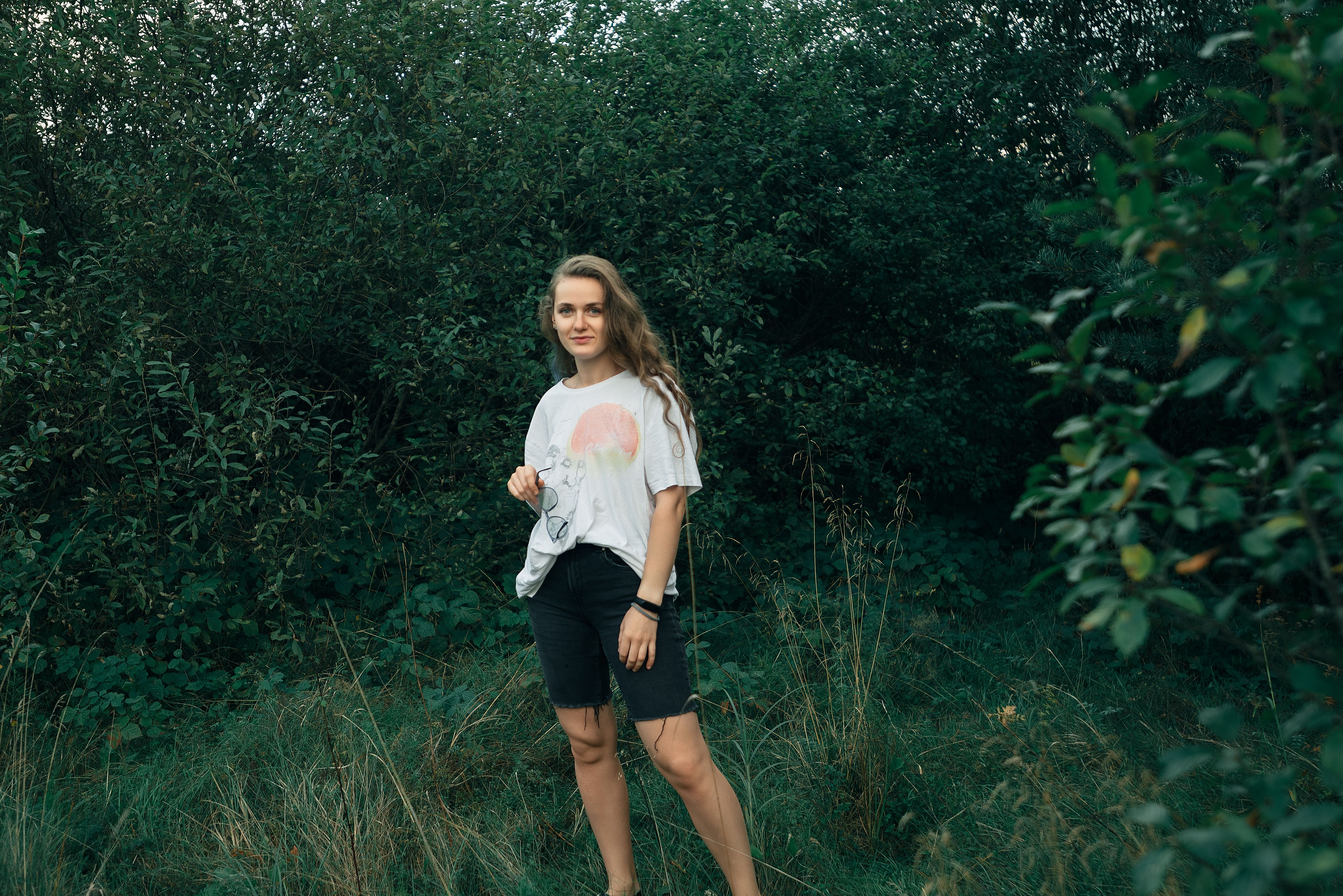A young woman stands confidently in lush outdoor greenery, dressed casually for a nature walk.