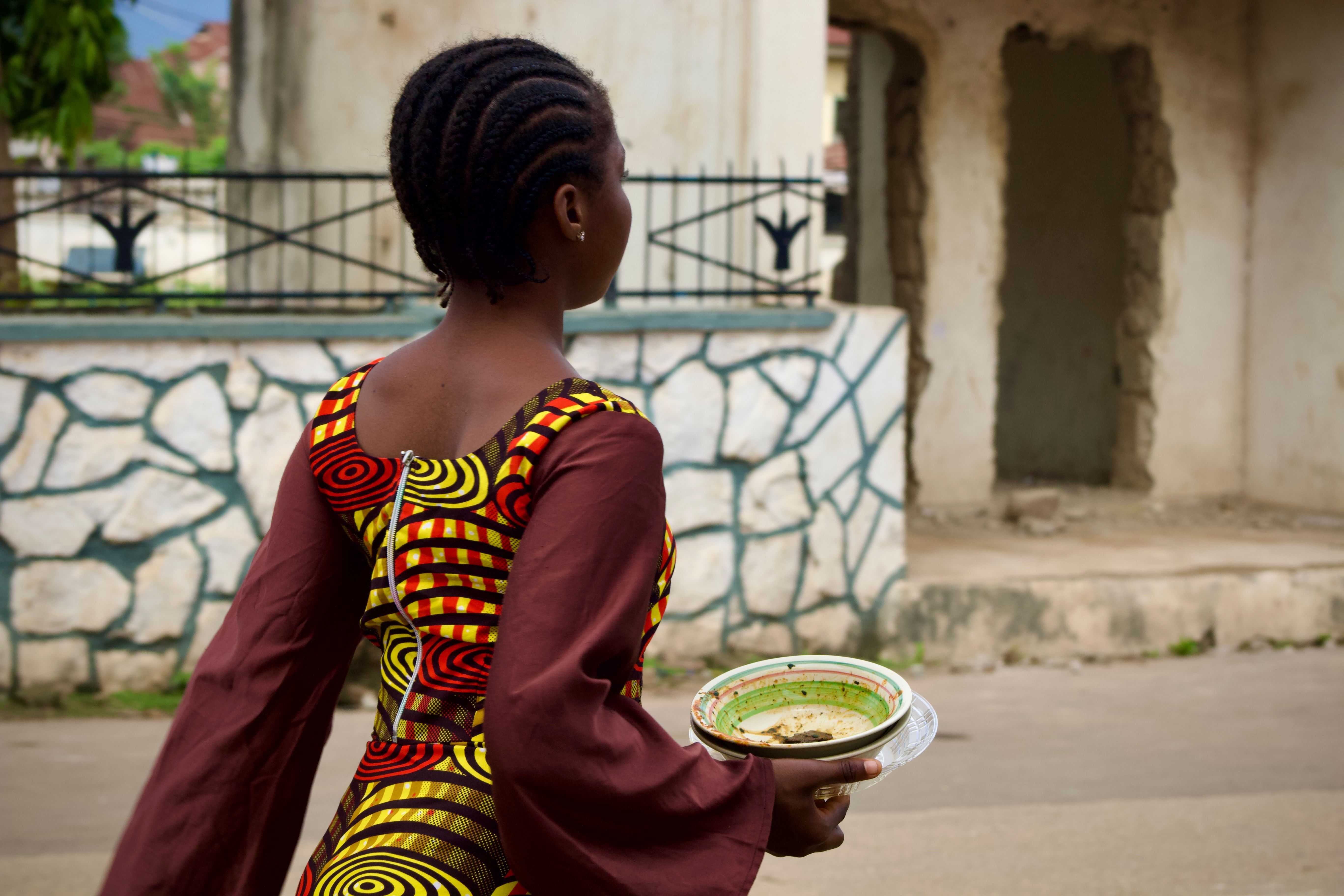 Back view of an African woman carrying a bowl while wearing colorful traditional attire outdoors.