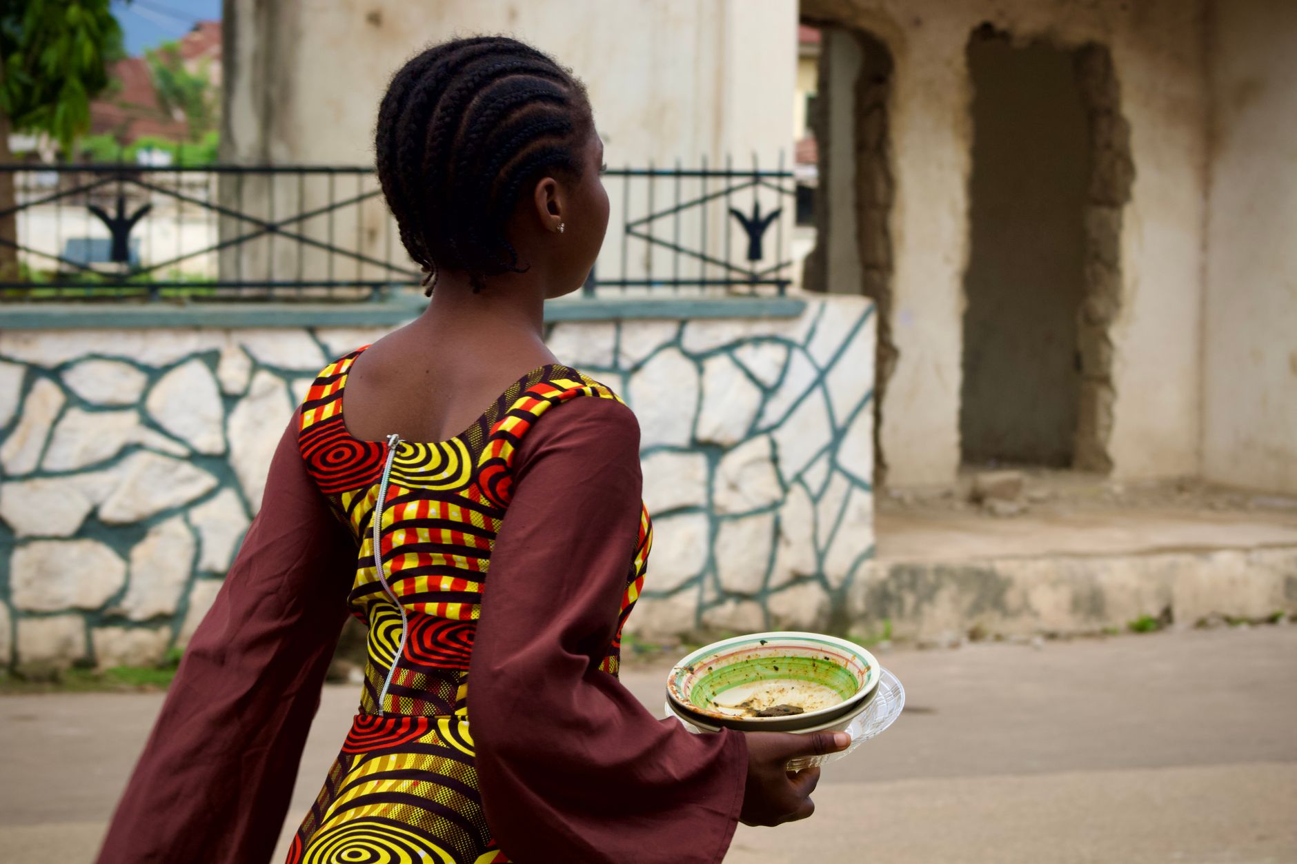 Back view of an African woman carrying a bowl while wearing colorful traditional attire outdoors.