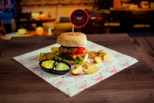 A juicy gourmet burger served with potato wedges and sauces in a cozy indoor setting, perfect for food lovers.