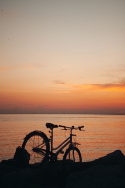 Free A lone bicycle silhouette rests on rocks by the sea during a warm, orange sunset. Stock Photo