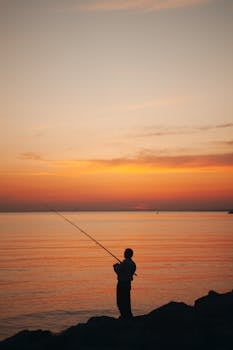 Silhouette of a person fishing by the ocean during a calming sunset.