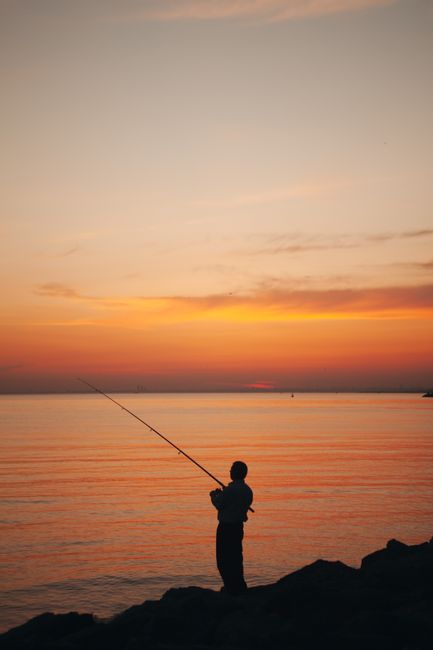 Silhouette of a person fishing by the ocean during a calming sunset.