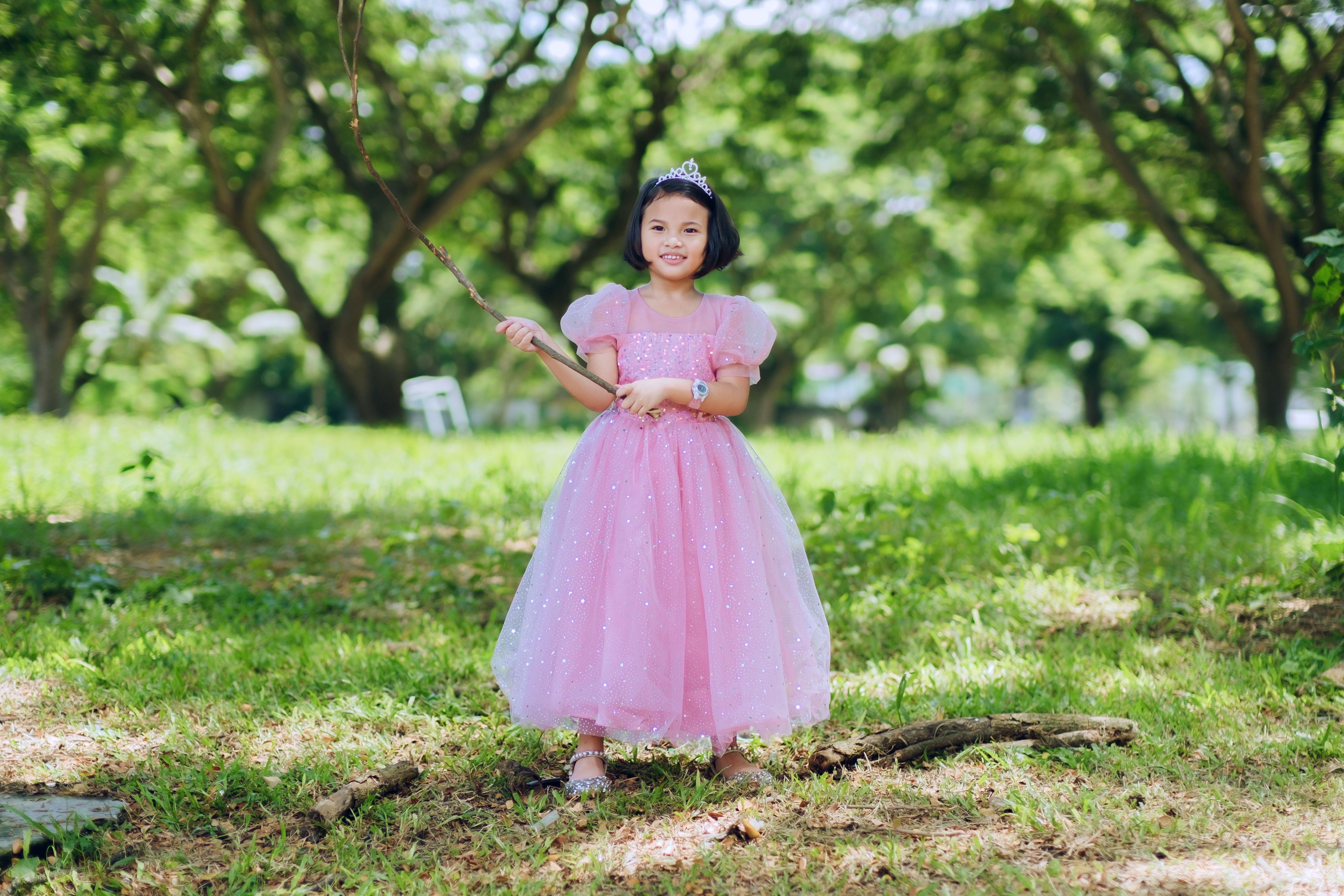 A young girl in a pink dress and tiara enjoying a playful moment in a lush green outdoor park.