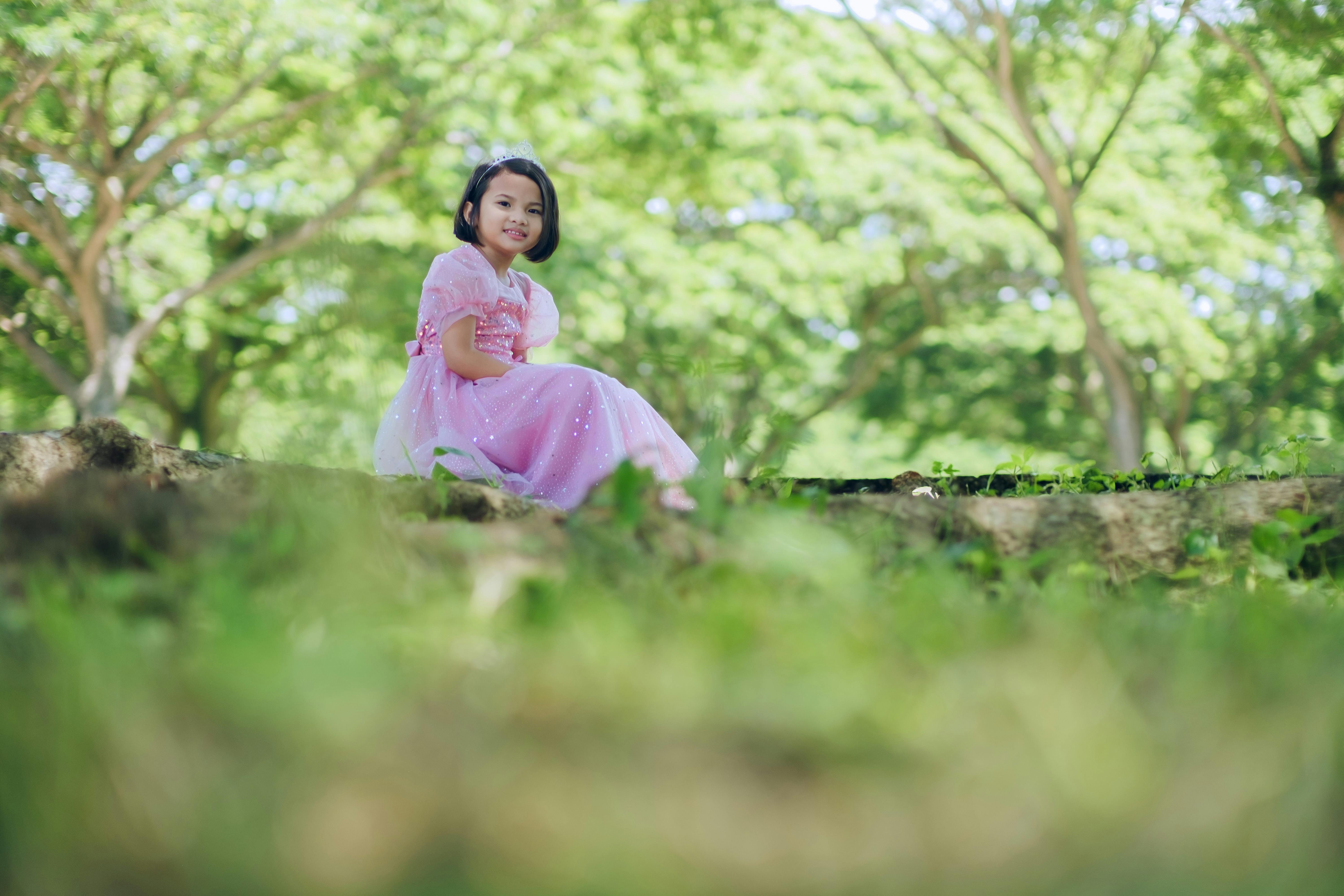 A young girl in a pink dress sits outdoors in a lush, green park setting.