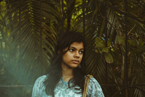 Foto De Retrato De Uma Mulher Com Flores Brancas E Azuis Posando Em Frente A Plantas De Folhas Verdes Olhando Para Longe